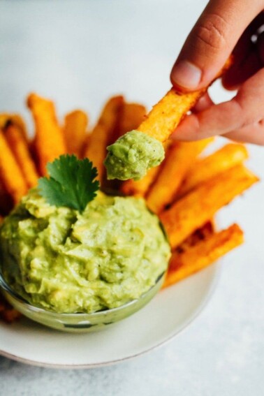 A hand using a jicama fry to scoop guacamole out of a bowl.