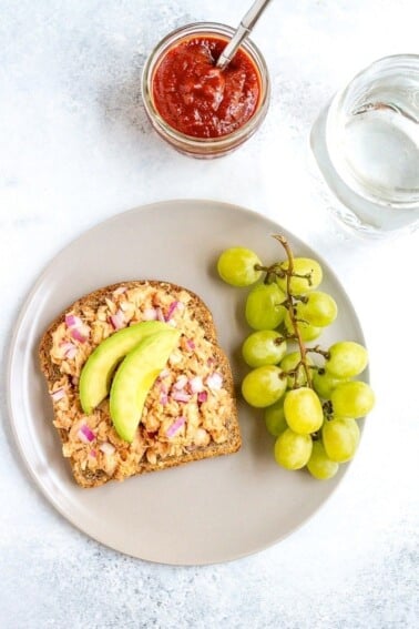 Tuna on bread topped with avocado with grapes on the side. Jam and water on the side.