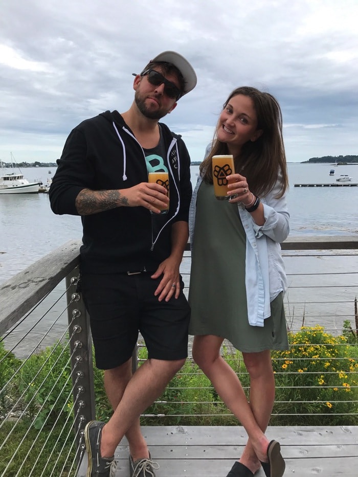 A woman and man smiling and posing with beer by the water.