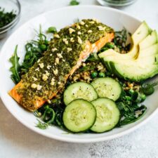 Pesto salmon, greens, herbs, cucumber, and avocado over rice on a white plate.