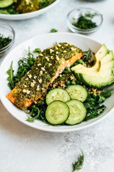 Pesto salmon, greens, herbs, cucumber, and avocado over rice on a white plate.