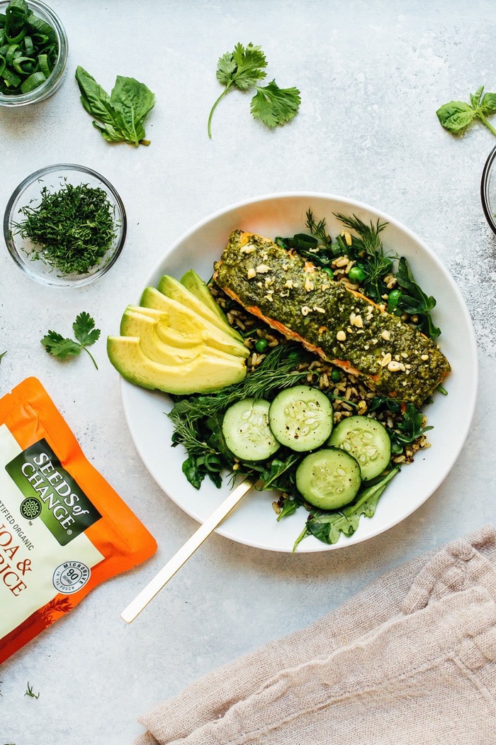 Pesto salmon, greens, herbs, cucumber, and avocado over rice, on a white plate surrounded by bowls of herbs.