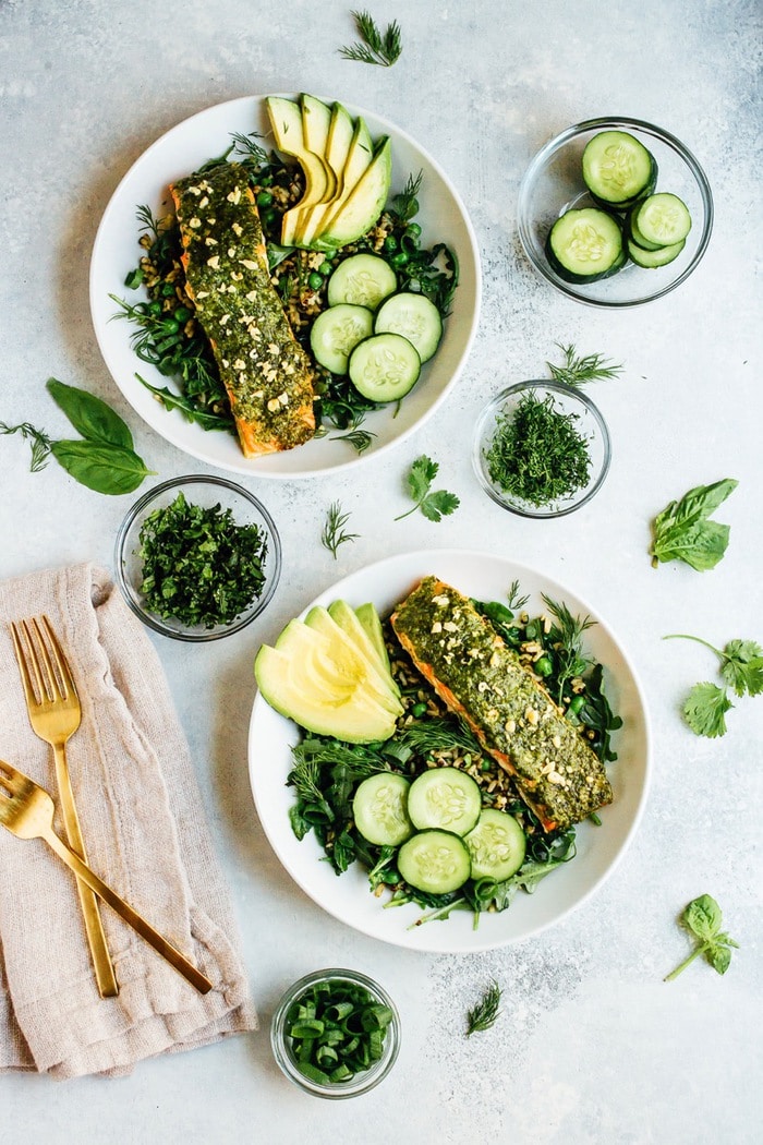 Two plates of pesto salmon, greens, herbs, cucumber, and avocado over rice, surrounded by bowls of herbs, forks and napkins.