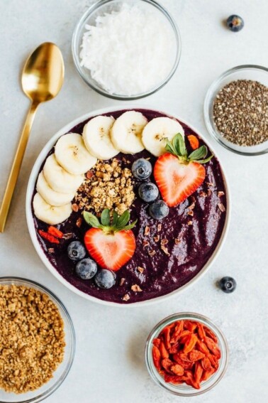 Maqui berry smoothie bowl topped with banana, strawberries, blueberries, and granola. Gold spoon laying beside the bowl along with bowls of dried berries, granola, coconut and chia.