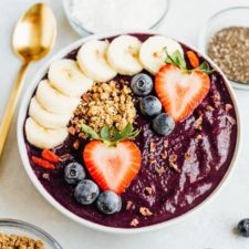 Maqui berry smoothie bowl topped with banana, strawberries, blueberries, and granola. Gold spoon laying beside the bowl along with bowls of dried berries, granola, coconut and chia.