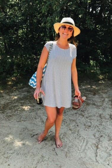 Woman smiling, wearing sunglasses and a short-sleeve striped sundress, standing in sand.