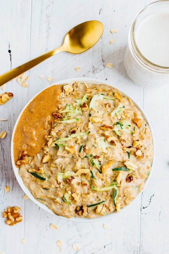 Bowl of zucchini bread oatmeal topped with walnuts and almond butter. Gold spoon and a glass of almond milk are next to the bowl.