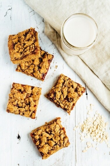 Peanut Butter and Jelly Crumble Bars on a table next to some oats and a glass of almond milk.