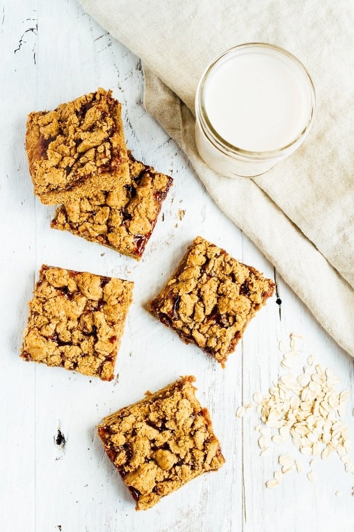 Peanut Butter and Jelly Crumble Bars on a table next to some oats and a glass of almond milk.