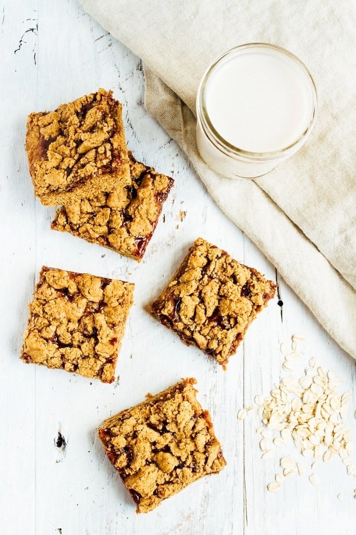 Peanut Butter and Jelly Crumble Bars Peanut Butter and Jelly Crumble Bars on a table next to some oats and a glass of almond milk.