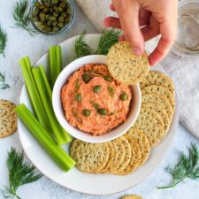 Hand scooping smoked salmon dip, off a plate with celery sticks and crackers.
