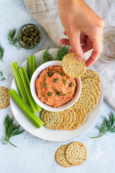 Hand scooping smoked salmon dip, off a plate with celery sticks and crackers.
