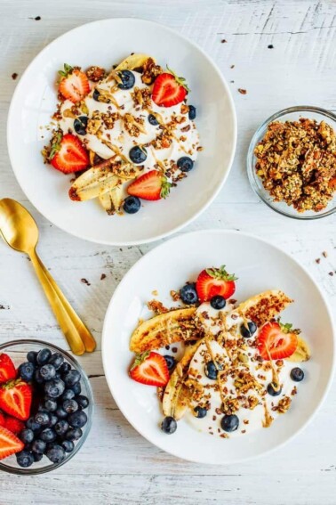 Two yogurt bowls with banana, strawberries, granola, and nut butter with a gold spoon, bowl of granola and bowl of strawberries and blueberries.