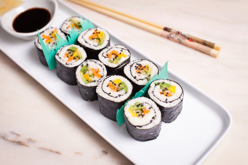 10 cauliflower rice sushi pieces on a white plate with soy sauce for dipping and chopsticks.