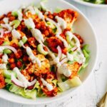 Buffalo cauliflower and chickpeas over lettuce with purple onion and dressing drizzled over in a white bowl on white wood.