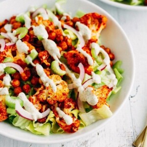 Buffalo cauliflower and chickpeas over lettuce with purple onion and dressing drizzled over in a white bowl on white wood.