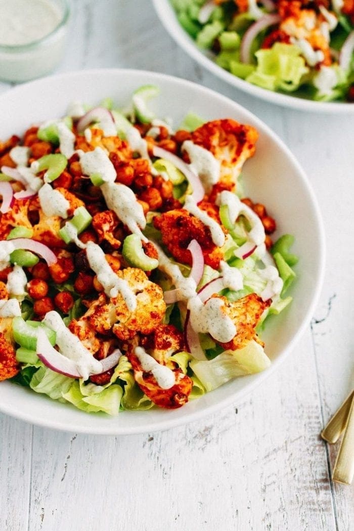 Buffalo cauliflower and chickpeas over lettuce with purple onion and dressing drizzled over in a white bowl on white wood.