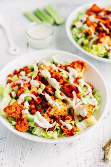 Two bowls of buffalo cauliflower and chickpeas over lettuce with purple onion and dressing drizzled on it. Dressing and celery sticks in the background.