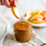 A hand dipping an apple into date caramel in a small jar.