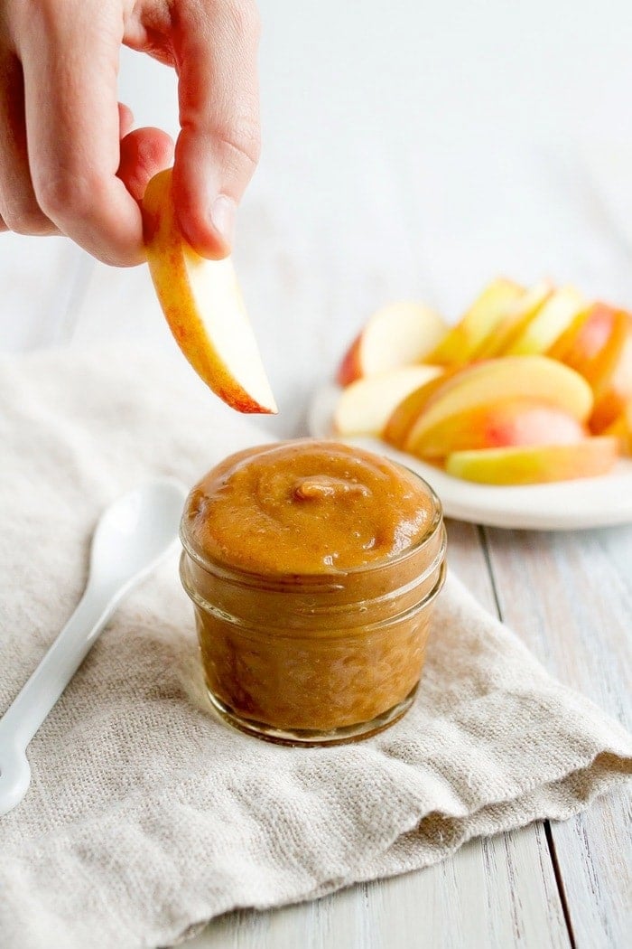 A hand holding an apple slice, about to dip into a glass jar of date caramel sauce.