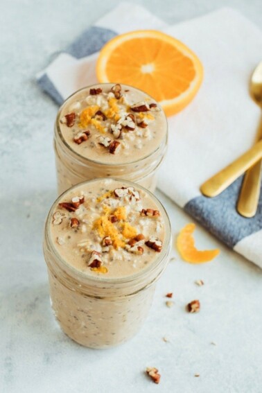 Two jars of orange vanilla overnight oats, topped with orange zest and pecans, with a slice of orange, and a towel next to them.