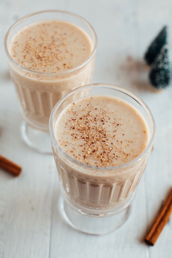 Healthy eggnog in glassed with nutmeg sprinkled on top. Mini Christmas trees and sticks of cinnamon sticks are in the background.