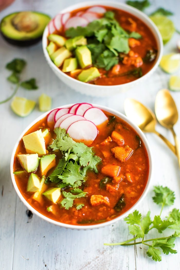 A close up of sweet potato and chicken soup topped with avocada, radishes, and cilantro. There is a second bowl out of focus in the background alongside two golden spoons.