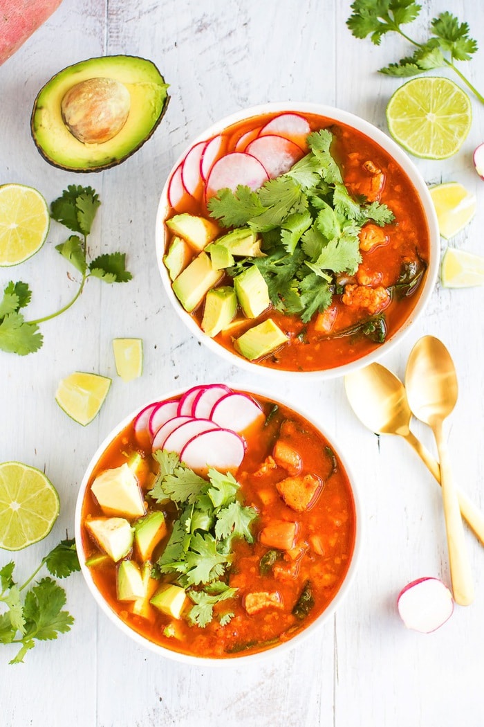 An overhead photo of two bowls filled with soup and topped with avocado, cilantro and radishes. There are limes, avocado, and radishes scattered around the bowl alongside two golden spoons.