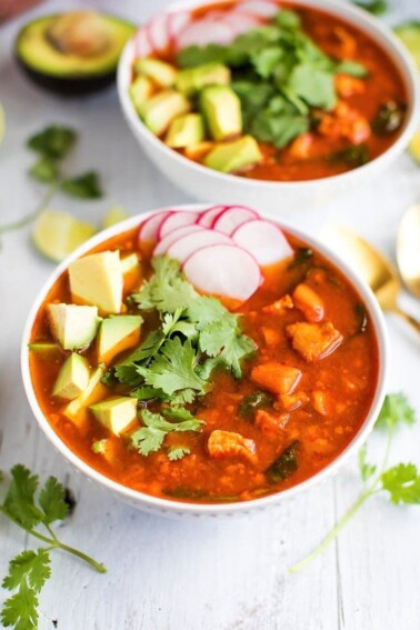 Two bowls of Mexican sweet potato chicken soup with avocado, radishes, and cilantro. Half an avocado, limes, and cilantro on the side.