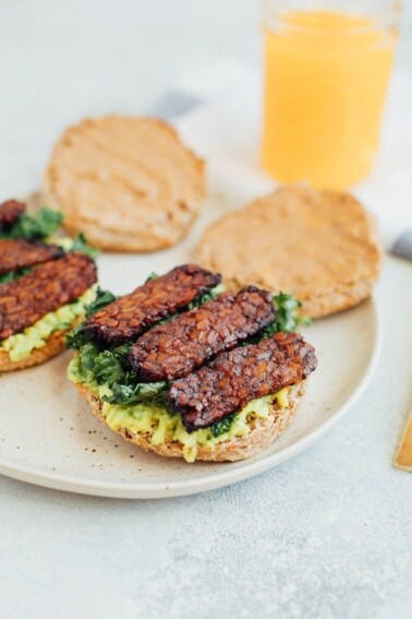 A vegan breakfast sandwich loaded with creamy avocado, almond butter, sautéed kale and tempeh bacon, on an english muffin. Orange juice in the background.