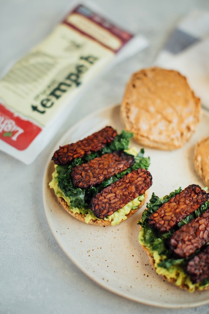 A vegan breakfast sandwich loaded with all the goods: creamy avocado, almond butter, sautéed kale and tempeh bacon, served on a perfectly toasted english muffin.