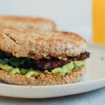 A vegan breakfast sandwich loaded with creamy avocado, almond butter, sautéed kale and tempeh bacon, on an english muffin.
