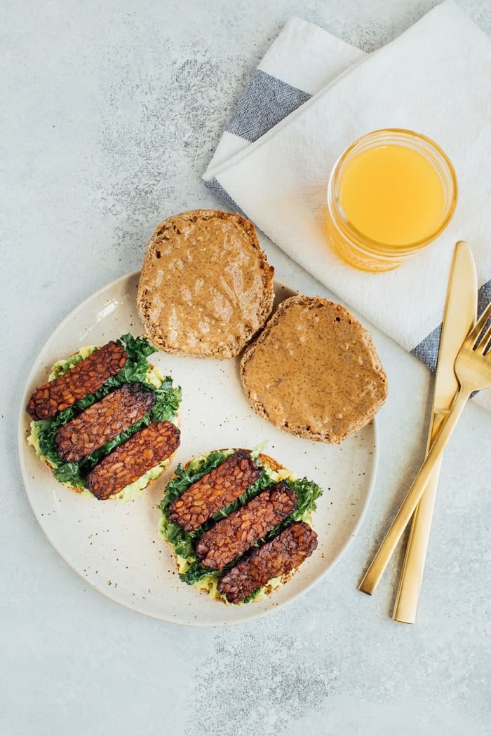 A vegan breakfast sandwich loaded with all the goods: creamy avocado, almond butter, sautéed kale and tempeh bacon, served on a perfectly toasted english muffin.