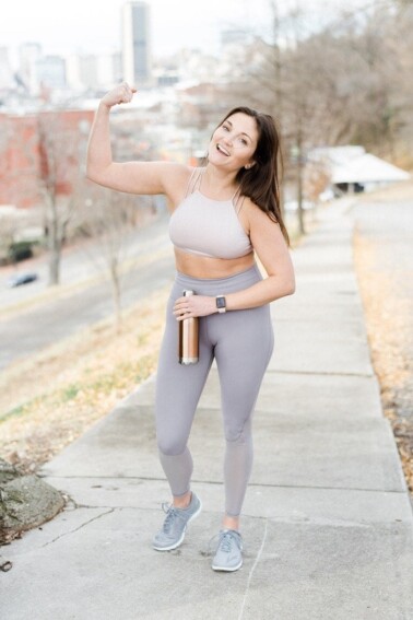 Woman flexing and smiling in athletic wear on a sidewalk.