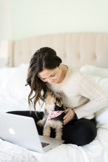 Woman in sweater and black pants on a bed playing with her yorkie and a Mac laptop.