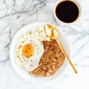 White bowl of oatmeal topped with almond butter, a fried egg with red pepper flakes, and scrambled egg whites. Black coffee next to the plate.