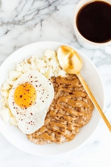 White bowl of oatmeal topped with almond butter, a fried egg with red pepper flakes, and scrambled egg whites. Black coffee next to the plate.