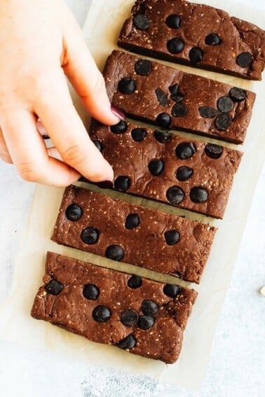 Hand grabbing a peanut butter chocolate protein bars with chocolate chips, from a row of bars on a white textured surface.