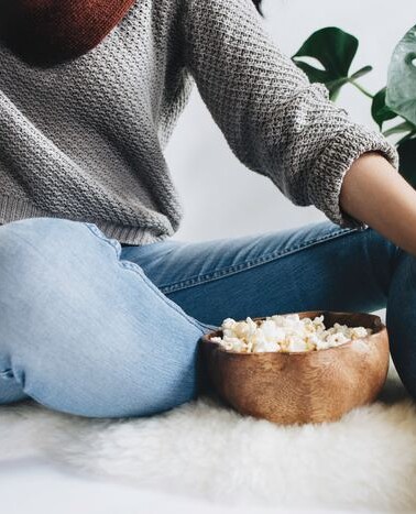 Woman sitting in jeans and a gray sweater with a bowl of popcorn.