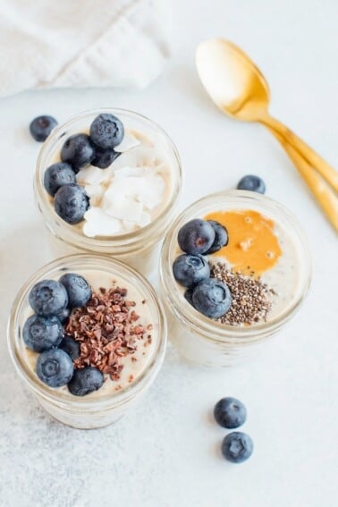Three pint-size mason jars with grain-free overnight "oats" and toppings. Two gold spoons on the right.