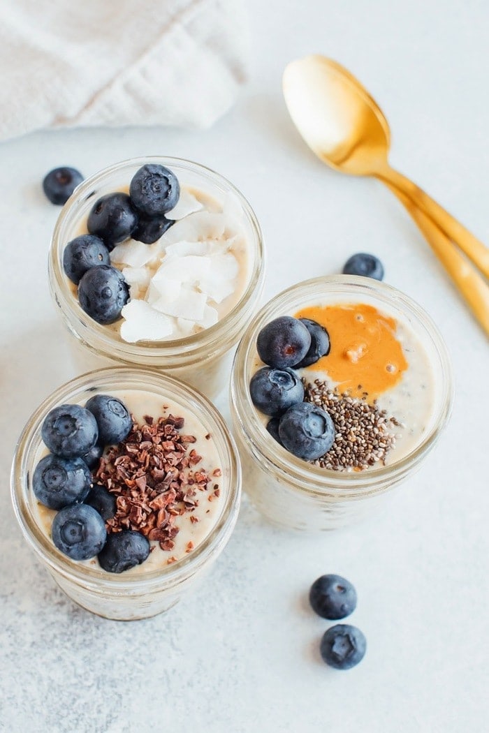 Three pint-size mason jars with grain-free overnight "oats" and toppings. Two gold spoons on the right. 