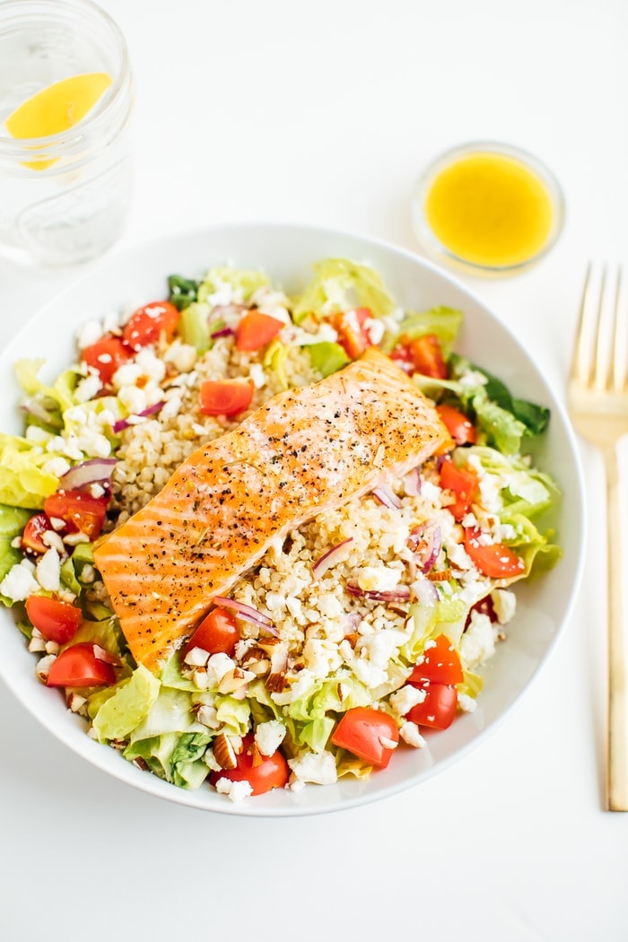 Salmon over Mediterranean salad in a white bowl with a fork and dressing on the side.