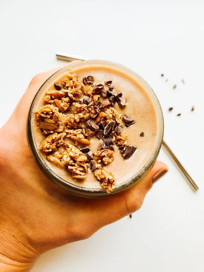 Overhead shot of a chocolate mint chip smoothie with granola in a mason jar being held by a women's hand with a stainless steel straw on the surface next to it. 