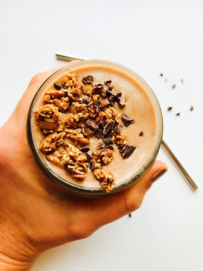 Overhead shot of a chocolate mint chip smoothie with granola in a mason jar being held by a women's hand with a stainless steel straw on the surface next to it. 