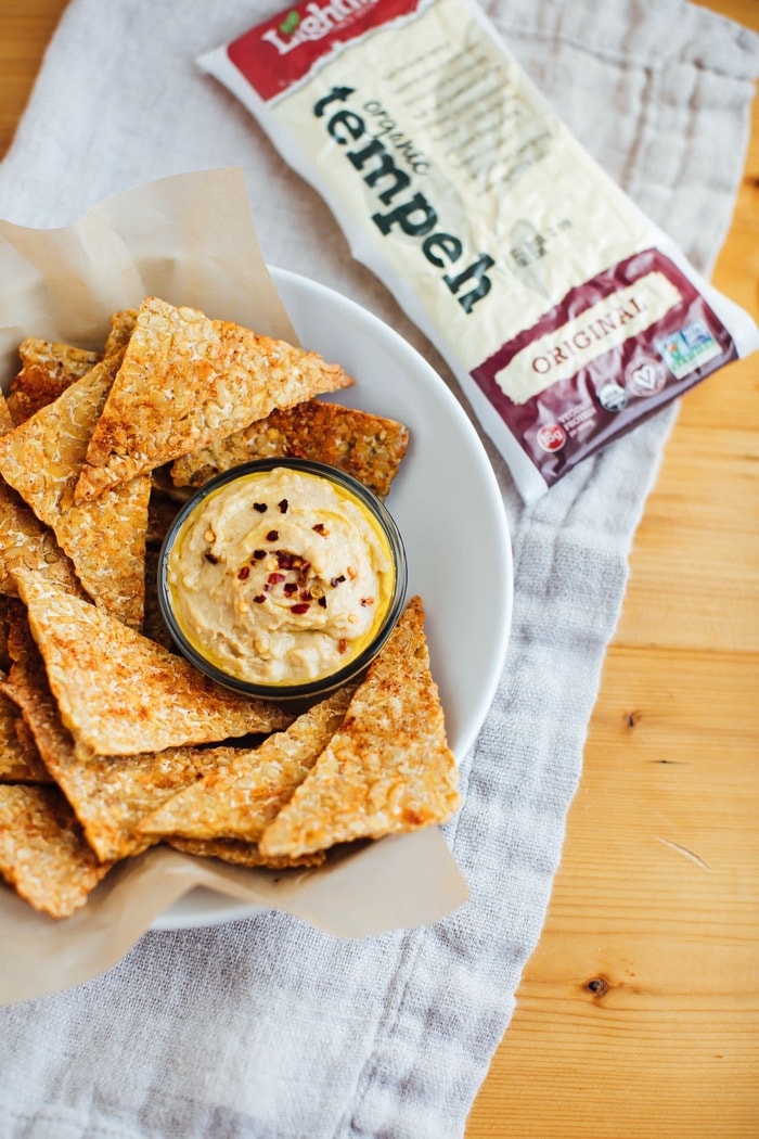 Close up of BBQ tempeh chips with hummus in a white bowl with brown parchment paper. Package of Lightlife tempeh in the background. 