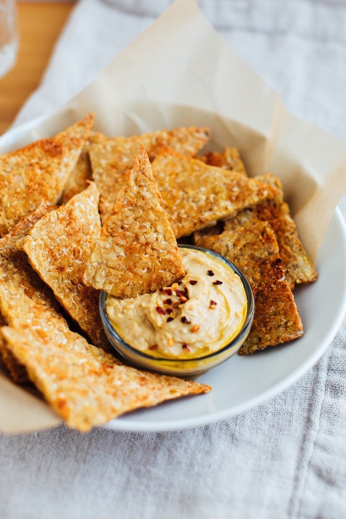 Baked tempeh chips in a white bowl with hummus. 