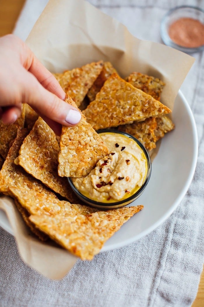 Hand with light pink nails holding a bbq baked tempeh chip and dipping it into hummus. 