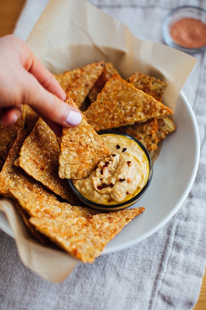 Hand with light pink nails holding a bbq baked tempeh chip and dipping it into hummus. 