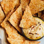 Baked bbq tempeh chips in a white bowl with hummus.