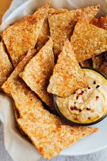 Baked bbq tempeh chips in a white bowl with hummus.